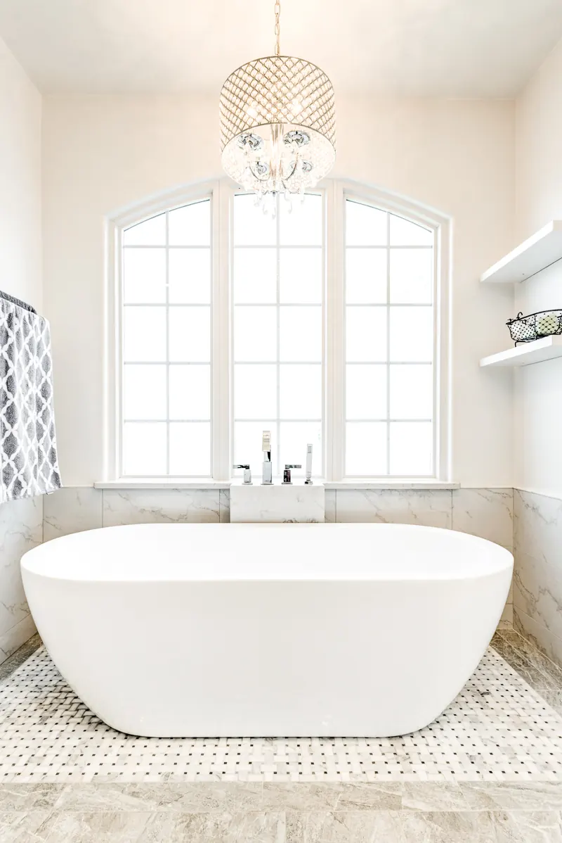 Freestanding tub under an arched window with a crystal chandelier and a marble mosaic floor, by Nomi